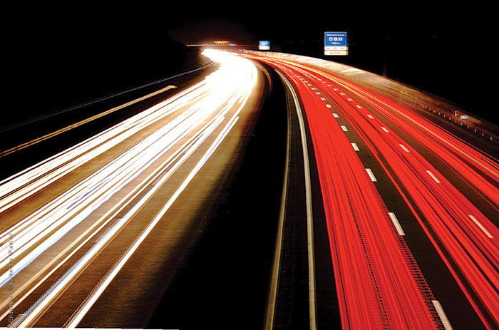 Das Bild zeigt eine Autobahn bei Nacht. Durch lange Belichtung des Photos sind links helle Lichtstreifen und rechts rote Lichtsstreifen auf den Fahrbahnen zu sehen.