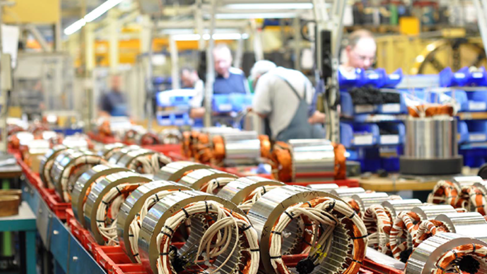 Das Bild zeigt eine große Anzahl mit Kabeln versehener Metallzylinder auf einem Fließband. Im Hintergrund arbeiten Menschen an verschiedenen Maschinen.