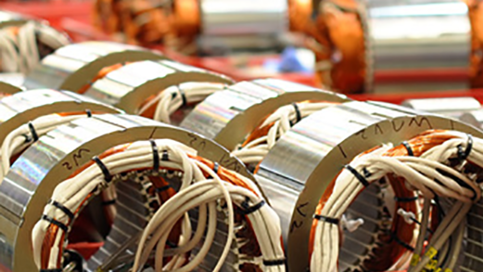 Das Bild zeigt mit Kabeln versehene Metallzylinder auf einem Fließband. Im Hintergrund arbeiten Menschen an verschiedenen Maschinen.
