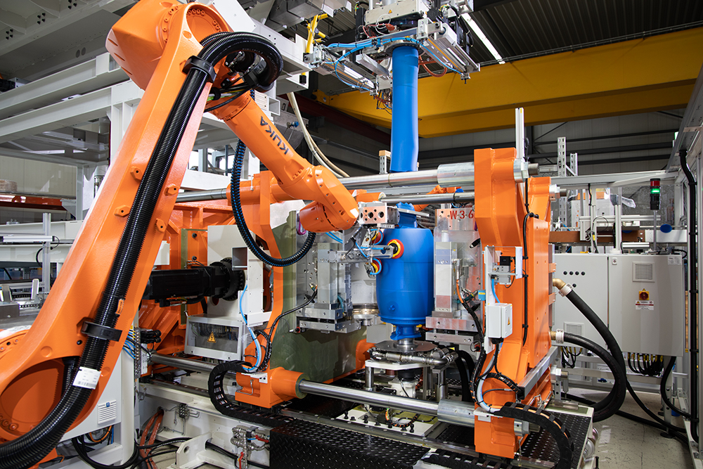 Das Foto zeigt orange Roboterarme, die nach einem blauen Kunststoff-Fass greifen. Das Fass wird aus gerade gegossen. Zu sehen ist ein Extruder am oberen Bildrand. Aus dem Extruder wird im Moment der Bildaufnahme ein blauer Kunststoff-Schlauch gepresst und in Form gebraucht. 