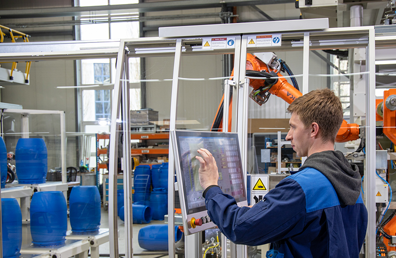 Ein junger Man steht in einer Fabrikhalle vor einem Bildschirm. Im Hintergrund sieht man einen orangen Roboterarm, der blaue Fässer anhebt.