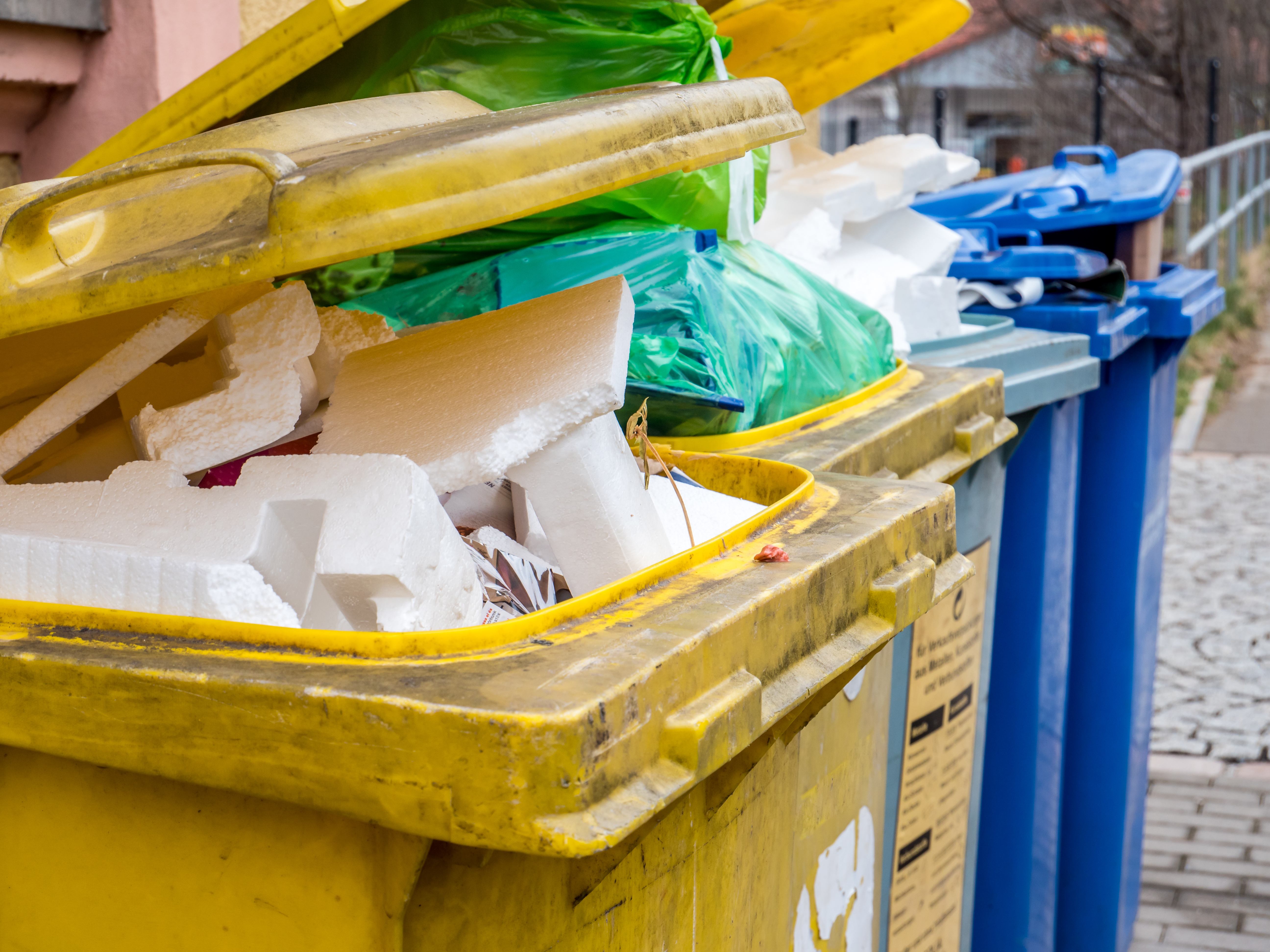 Das Bild zeigt gelbe und blaue Mülltonnen, die mit Plastik- und Styroporabfällen gefüllt sind.