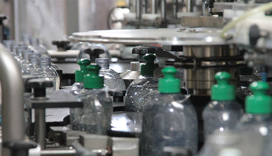 Das Bild zeigt in Nahaufnahme ein Förderband, auf dem Plastikflaschen für Spülmittel aufgereiht transportiert werden. Die Flaschen sind durchsichtig und haben einen grünen Verschluss.