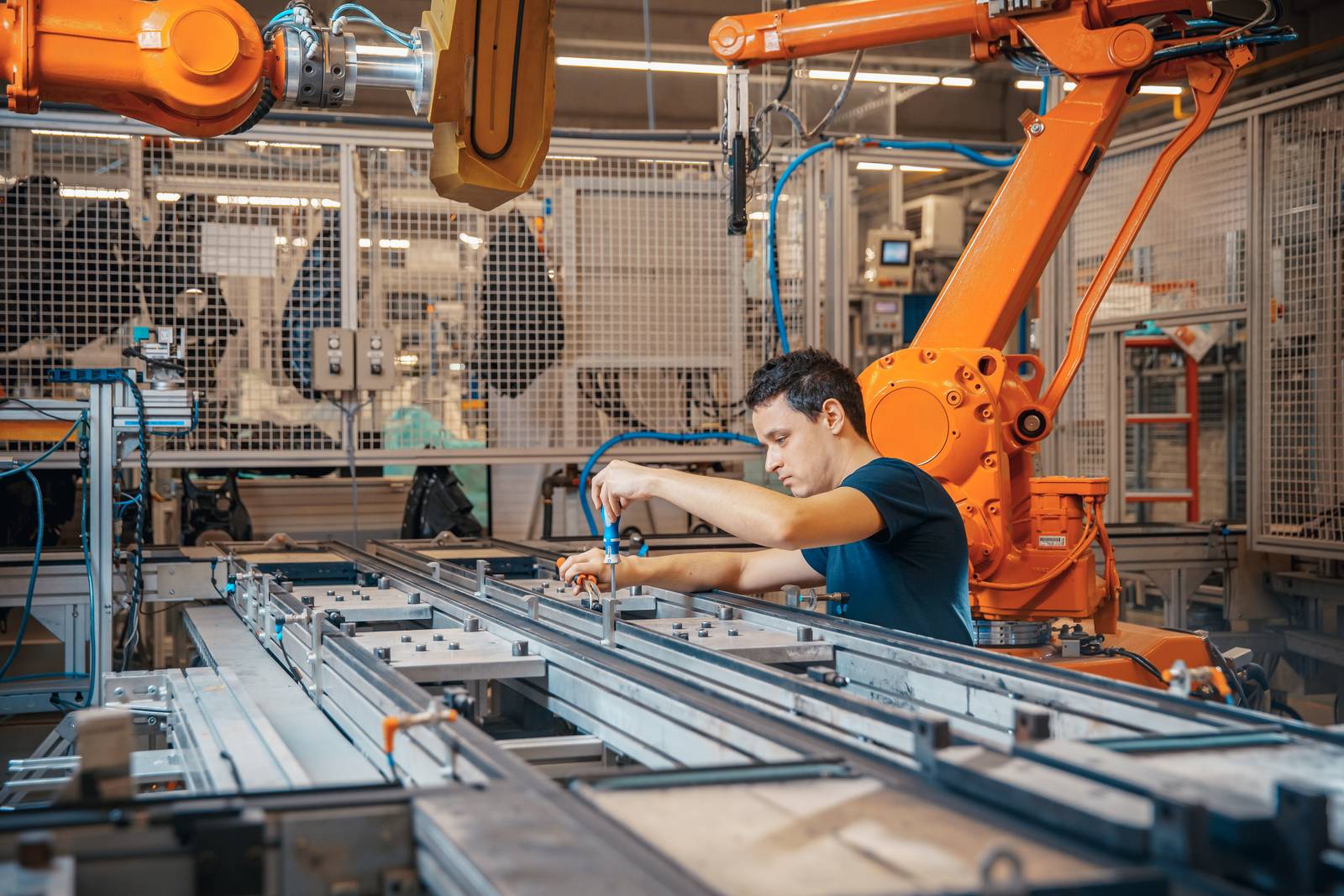 Ein Mann steht mit einem blauen Schraubendreher in der Hand an einem Montagetisch. Im Hintergrund sind orangefarbene Roboterarme zu sehen.
