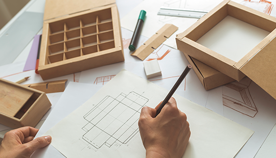 Das Bild zeigt eine Hand, die Produktentwürfe einer Schachtel zeichnet. Die Vorlage für die Schachtel ist neben Bleistiften, Linealen und Kartons ebenfalls im Bild zu sehen.