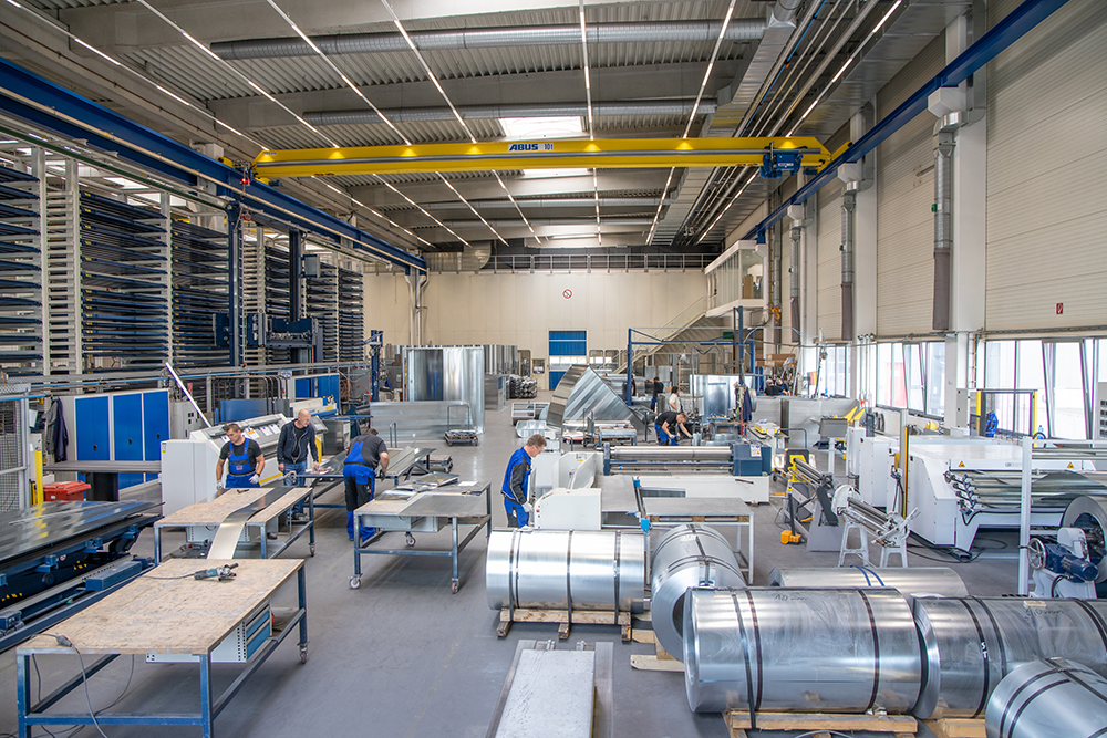 Blick in eine Produktionsstätte aus dem Metallbau. In der Halle sind verschiedene Mitarbeitende an verschiedene Werktischen zu sehen. 