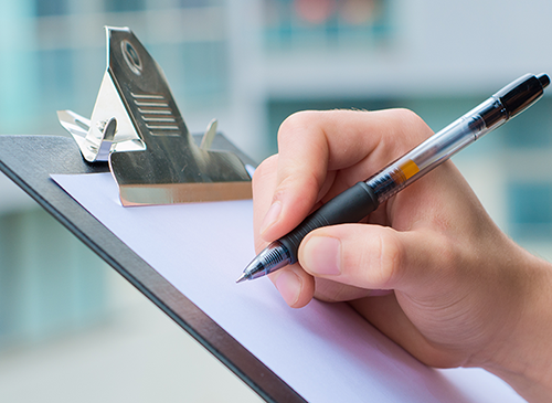 Detailaufnahme einer Person, die einen Stift in der Hand hält und damit auf ein Klemmbrett schreibt.
