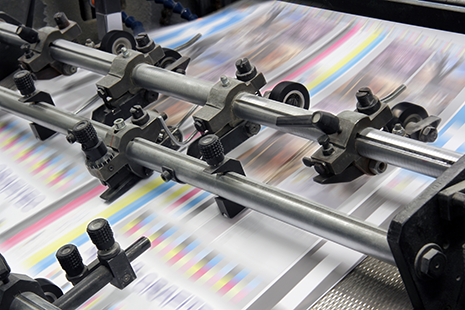 Das Bild zeigt eine Momentaufnahme einer professionellen Druckmaschine, während das Papier in den Farben Cyan, Magenta, Yellow und Key (schwarz) bedruckt wird.